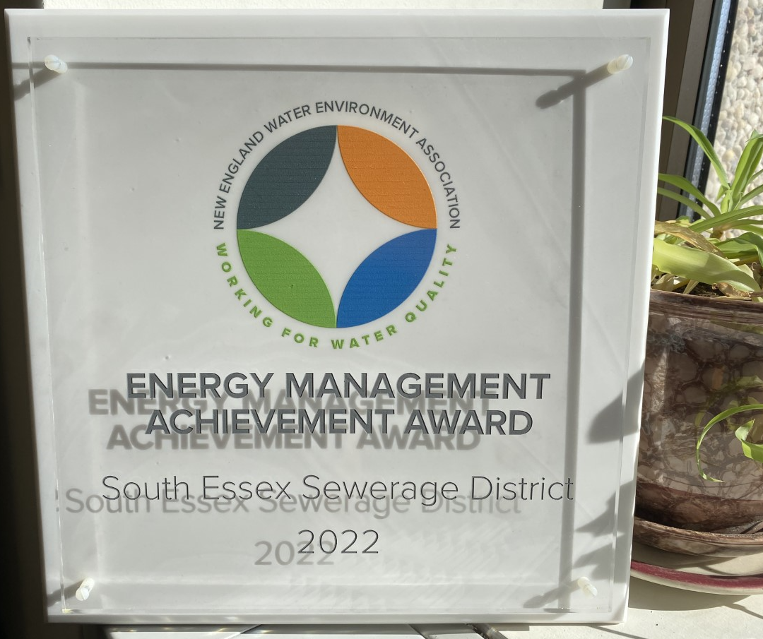 This is a photo of the 2022 New England Water Environment Association Energy Management Achievement Award. It is made of glass, with a solid white base and a clear glass top. The NEWEA logo is printed on the white base. It is a logo consisting of a white circle with gray, orange, green, and blue semicircles facing the center of it. It has the organization's name spelled around it in gray, and underneath curving up, the phrase, "Working for Water Quality" in green. On the clear glass is printed the award name, the District's name, and the year 2022. You can see all of this at once due to the layered glass. This was awarded to the South Essex Sewerage District in recognition of ten years of energy management and environmental stewardship.