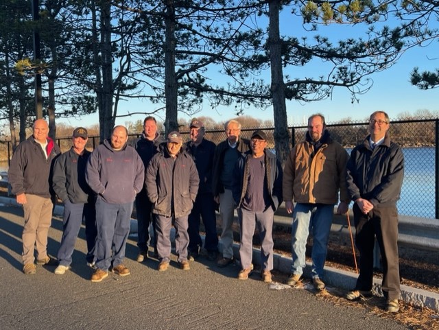 In this photo, ten men stand in front of Salem Harbor along a black, chain link fence. They are all employees of the South Essex Sewerage District and are gathered in celebration of winning the 2022 New England Water Environment Association Energy Management Award. They stand in a semicircle, smiling in the light of the setting sun.
