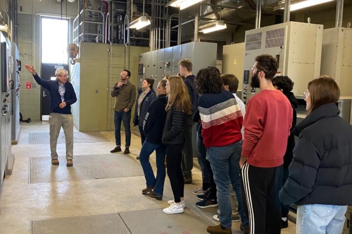 This is a photo of an Endicott College class on a tour of the Influent Building at South Essex Sewerage District. District Engineer, Mike Wilson, leads the discussion as students and professor look around the large, blinking control panels in front of them.