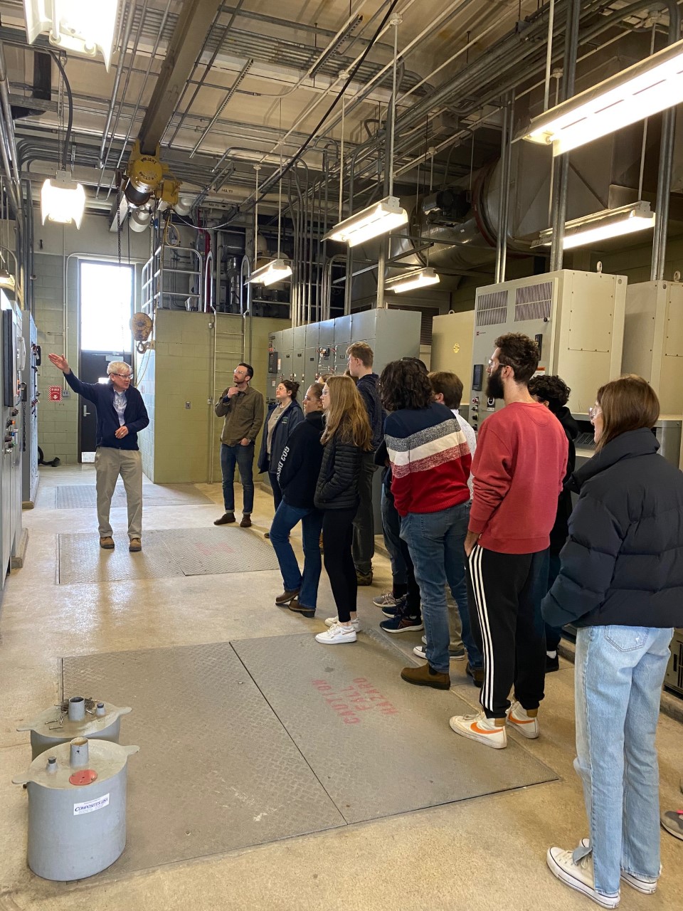 This is a photo of an Endicott College class on a tour of the Influent Building at South Essex Sewerage District. District Engineer, Mike Wilson, leads the discussion as students and professor look around the large, blinking control panels in front of them.