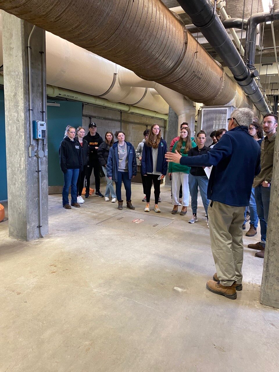 This is a photo of an Endicott College class on a tour of the Effluent Building at South Essex Sewerage District. District Engineer, Mike Wilson, leads the discussion as students and professor look around a large room full of enormous pipes.