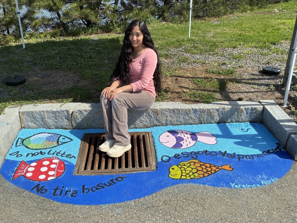 Photo of Salem high school student, Yareimee Abreu, sitting next to the RV Station Storm Drain mural she painted with Salem Sound Coastwatch's Susan Marsh. It features an ocean background with colorful fish, and says, "Do not litter", "No tire basura", "O esgoto vai para ocean" with the SSCW logo in the top right corner.