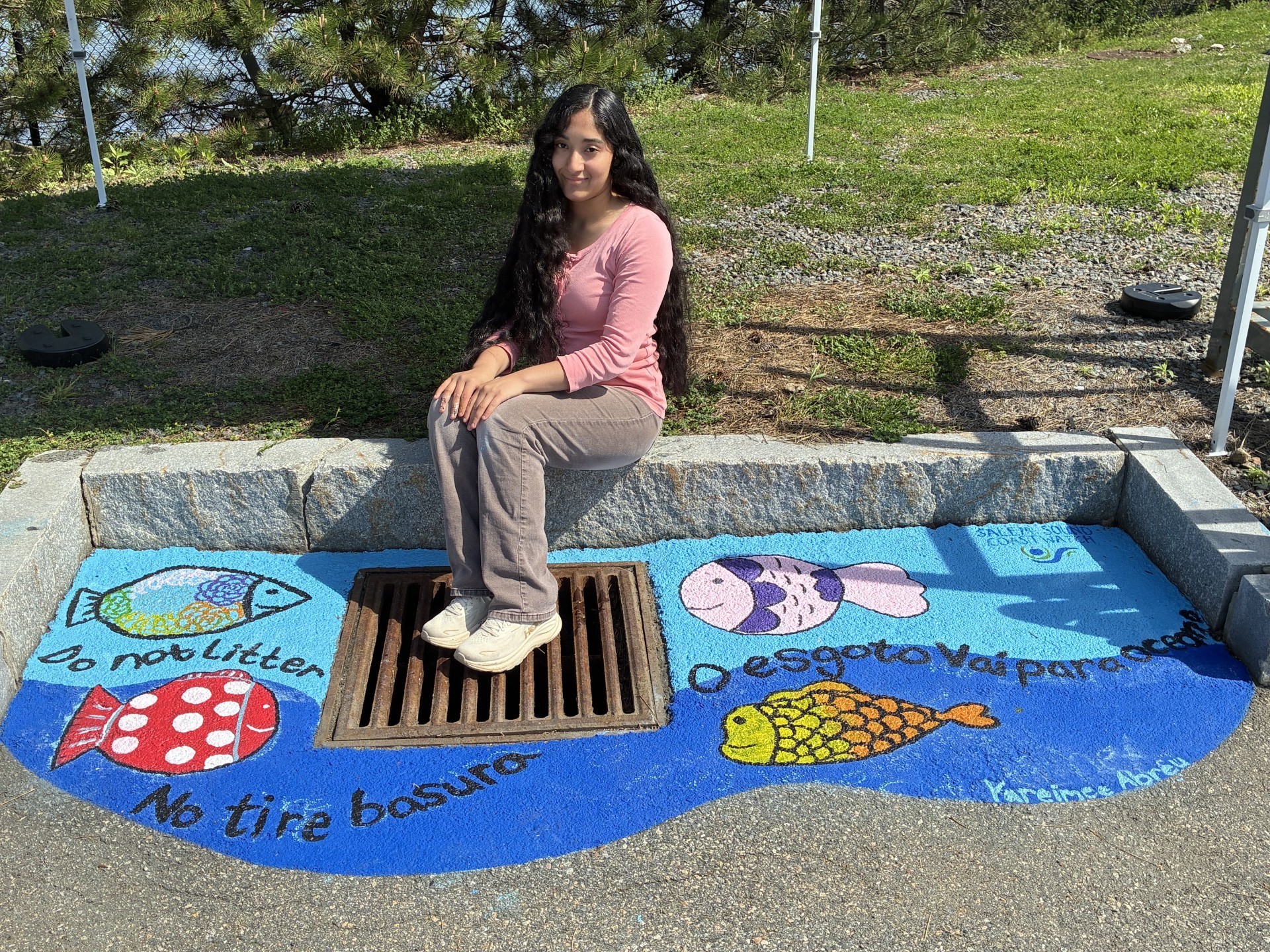 Photo of Salem high school student, Yareimee Abreu, sitting next to the RV Station Storm Drain mural she painted with Salem Sound Coastwatch's Susan Marsh. It features an ocean background with colorful fish, and says, "Do not litter", "No tire basura", "O esgoto vai para ocean" with the SSCW logo in the top right corner.